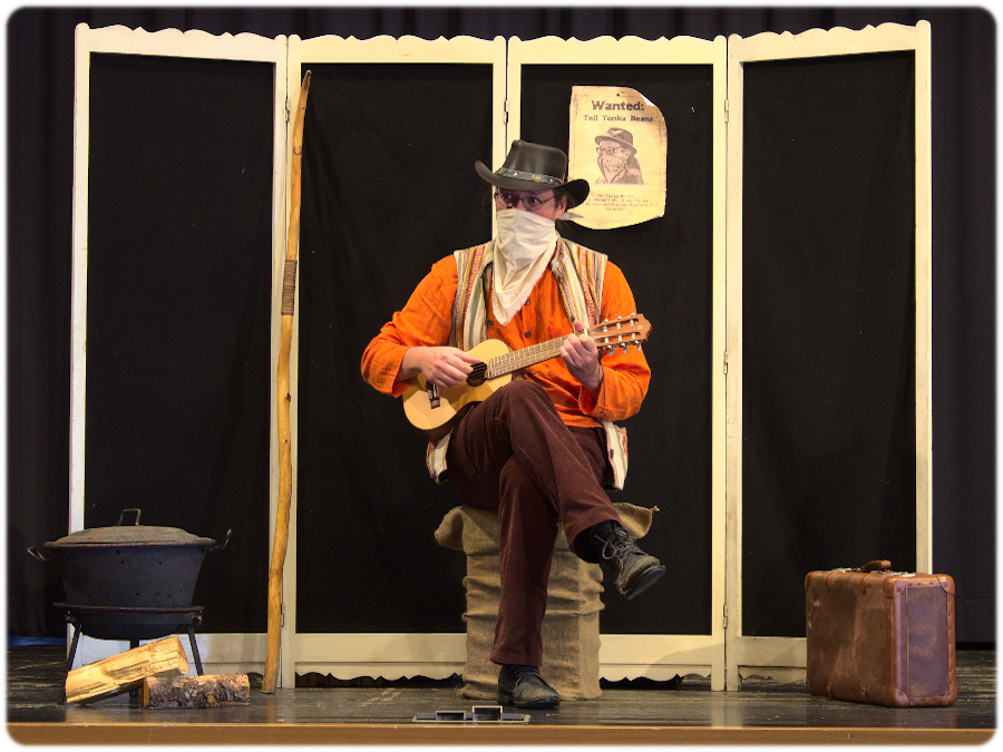 Ein verkleideter Darsteller in einem Cowboy-Outfit spielt auf einer Gitarre, während er auf einem Sack sitzt. Im Hintergrund sind eine alte Koffer und ein Kochtopf zu sehen, sowie ein 'Wanted'-Plakat.